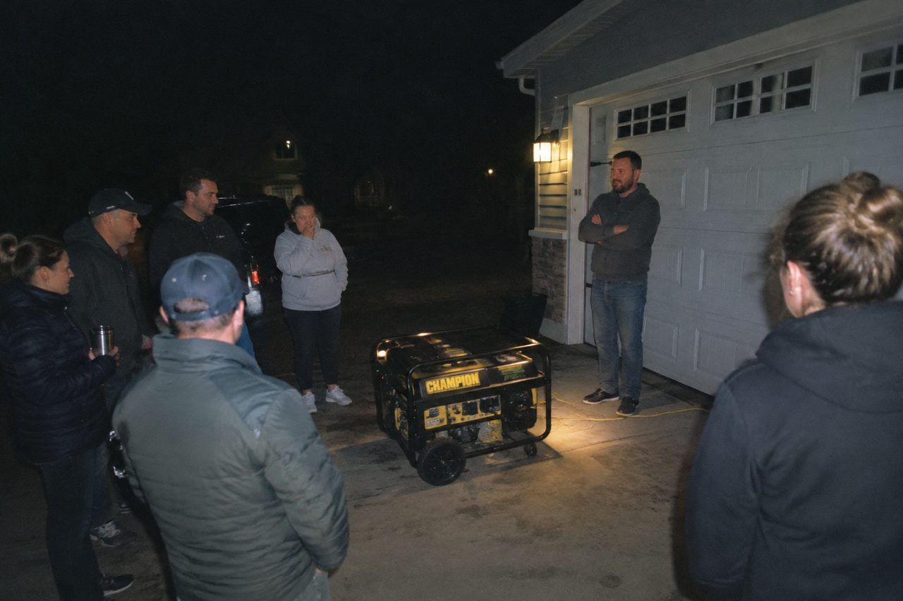 A crowd of neighbours gathered at night around a running generator in a suburban driveway, the homeowner standing in front of his garage