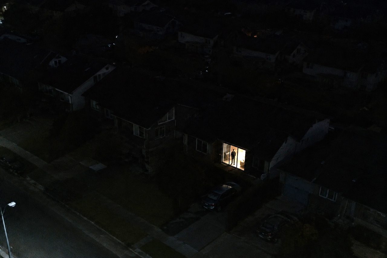 Drone view over a dark suburb at night, every house black except one lit window with a man standing in the doorway
