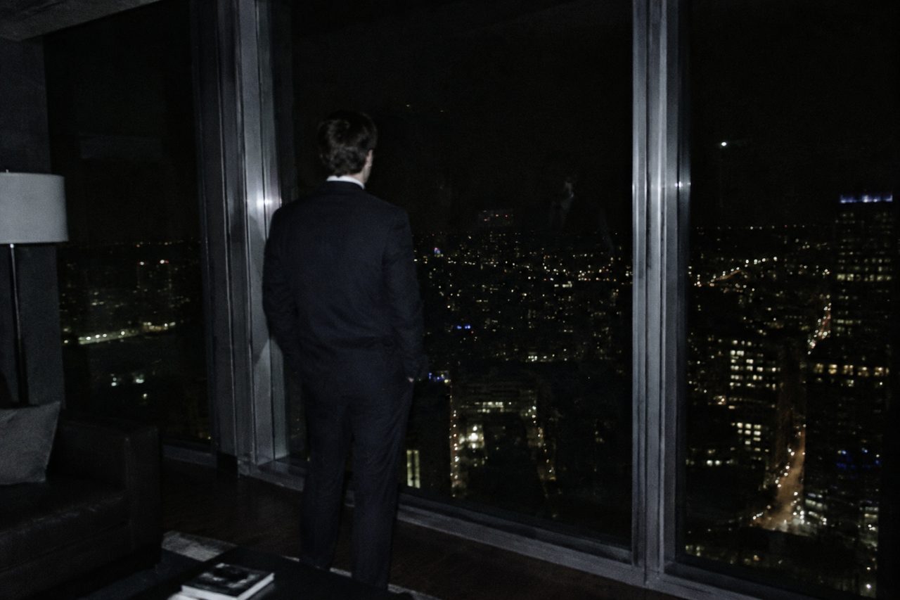 A man seen from behind at a dark high-rise window at night, the city lights spread out below