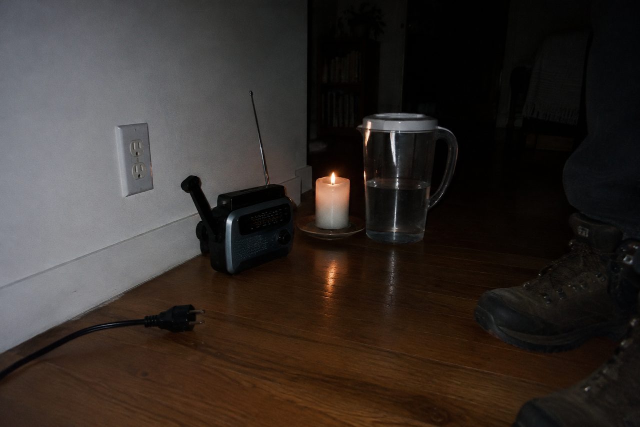 An unplugged power cord on a wooden floor beside a hand-crank radio, a candle and a jug of water
